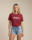 Woman wearing a maroon t-shirt with 'Montavor' logo and denim shorts on a beige background