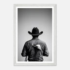 Black and white photograph of a rodeo cowboy viewed from behind wearing a western hat with a numbered rodeo tag pinned to his shirt after competition.