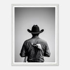 Black and white photograph of a rodeo cowboy viewed from behind wearing a western hat with a numbered rodeo tag pinned to his shirt after competition.
