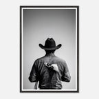 Black and white photograph of a rodeo cowboy viewed from behind wearing a western hat with a numbered rodeo tag pinned to his shirt after competition.