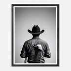 Black and white photograph of a rodeo cowboy viewed from behind wearing a western hat with a numbered rodeo tag pinned to his shirt after competition.