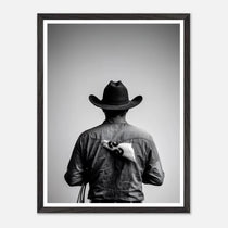 Black and white photograph of a rodeo cowboy viewed from behind wearing a western hat with a numbered rodeo tag pinned to his shirt after competition.