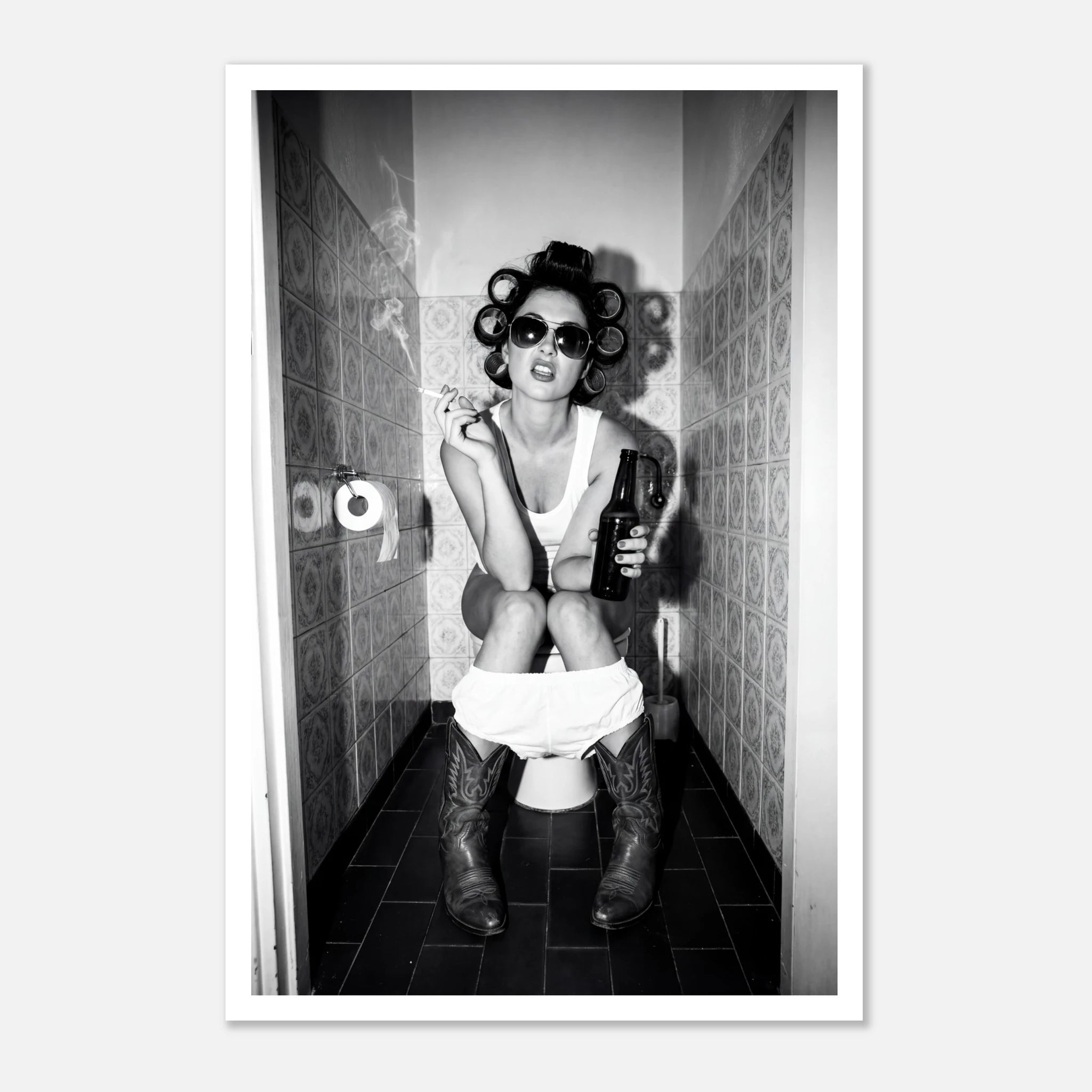 Black and white photograph of a rebellious cowgirl wearing cowboy boots and sunglasses sitting in a tiled restroom holding a cigarette and beer.