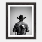 Black and white photograph of a rodeo cowboy viewed from behind wearing a western hat with a numbered rodeo tag pinned to his shirt after competition.