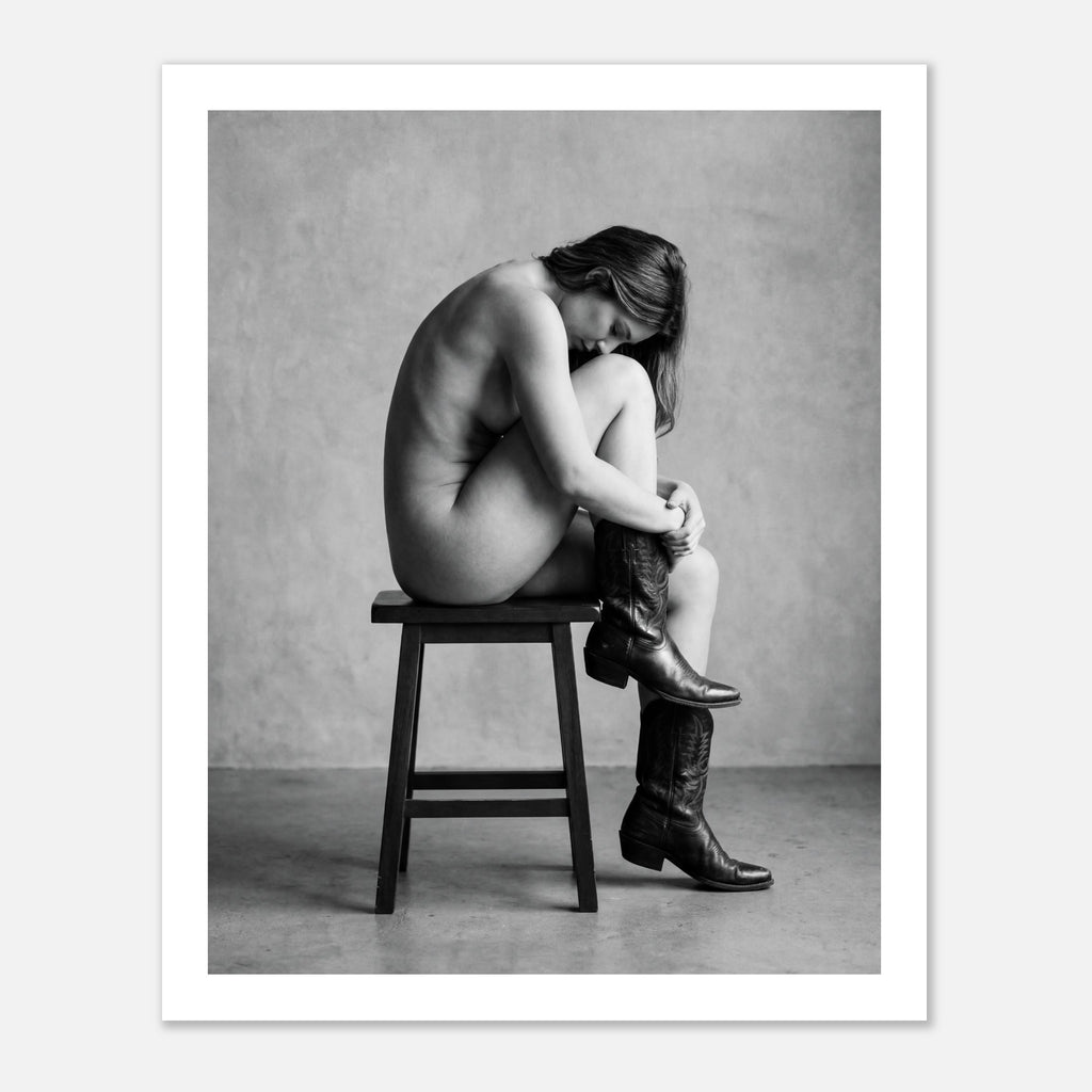Black and white fine art photograph of a woman seated on a stool wearing cowboy boots in a minimalist studio setting.