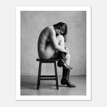 Black and white fine art photograph of a woman seated on a stool wearing cowboy boots in a minimalist studio setting.