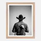Black and white photograph of a rodeo cowboy viewed from behind wearing a western hat with a numbered rodeo tag pinned to his shirt after competition.