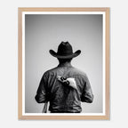 Black and white photograph of a rodeo cowboy viewed from behind wearing a western hat with a numbered rodeo tag pinned to his shirt after competition.