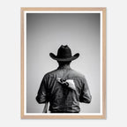 Black and white photograph of a rodeo cowboy viewed from behind wearing a western hat with a numbered rodeo tag pinned to his shirt after competition.