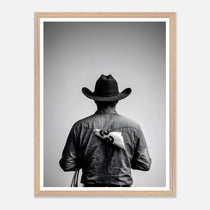 Black and white photograph of a rodeo cowboy viewed from behind wearing a western hat with a numbered rodeo tag pinned to his shirt after competition.