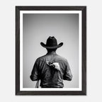 Black and white photograph of a rodeo cowboy viewed from behind wearing a western hat with a numbered rodeo tag pinned to his shirt after competition.