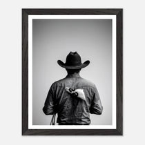 Black and white photograph of a rodeo cowboy viewed from behind wearing a western hat with a numbered rodeo tag pinned to his shirt after competition.