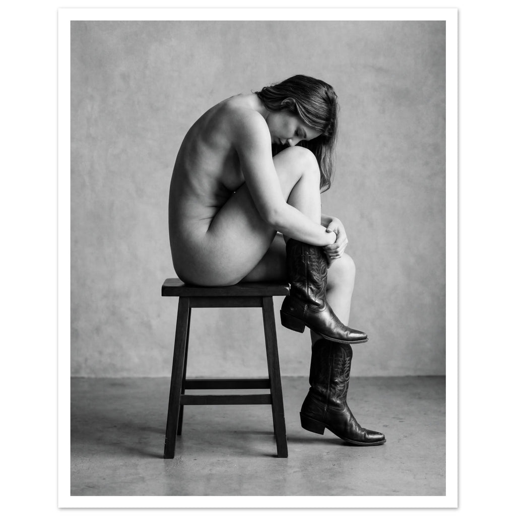 Black and white fine art photograph of a woman seated on a stool wearing cowboy boots in a minimalist studio setting.