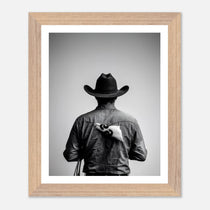 Black and white photograph of a rodeo cowboy viewed from behind wearing a western hat with a numbered rodeo tag pinned to his shirt after competition.