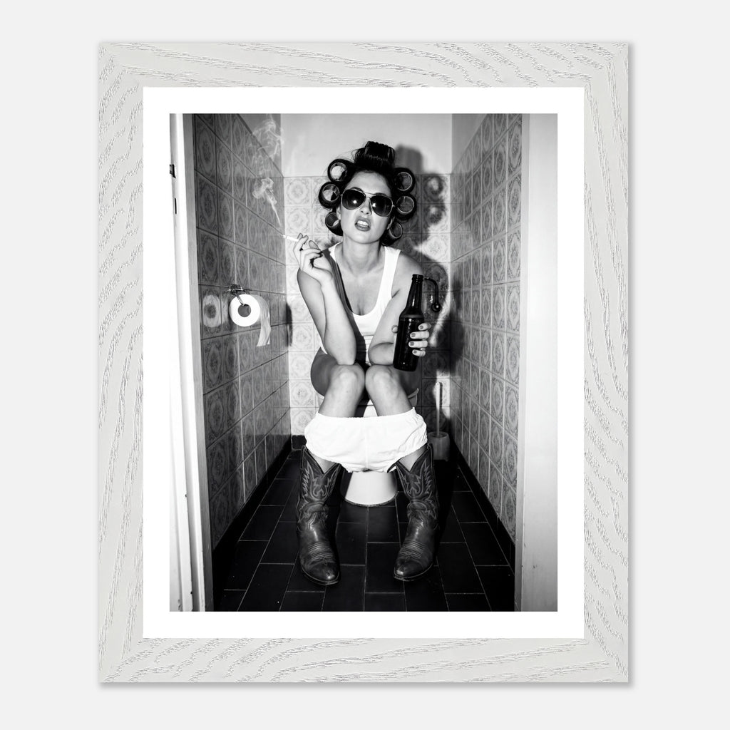 Black and white photograph of a rebellious cowgirl wearing cowboy boots and sunglasses sitting in a tiled restroom holding a cigarette and beer.