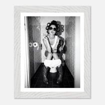 Black and white photograph of a rebellious cowgirl wearing cowboy boots and sunglasses sitting in a tiled restroom holding a cigarette and beer.