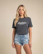 Woman wearing a dark gray t-shirt with 'Montavor Western Wear' text and denim shorts on a beige background