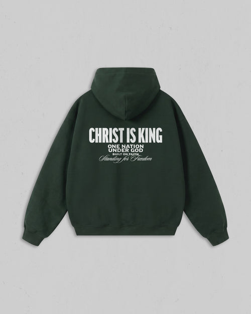 A dark forest green hoodie shown from the back, featuring white printed text that reads 'CHRIST IS KING' with smaller text below reading 'ONE NATION UNDER GOD' and 'Standing for Freedom'. The hoodie is displayed flat against a neutral light gray background.