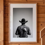 Black and white photograph of a rodeo cowboy viewed from behind wearing a western hat with a numbered rodeo tag pinned to his shirt after competition.