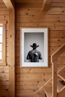 Black and white photograph of a rodeo cowboy viewed from behind wearing a western hat with a numbered rodeo tag pinned to his shirt after competition.