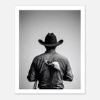 Black and white photograph of a rodeo cowboy viewed from behind wearing a western hat with a numbered rodeo tag pinned to his shirt after competition.