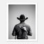 Black and white photograph of a rodeo cowboy viewed from behind wearing a western hat with a numbered rodeo tag pinned to his shirt after competition.