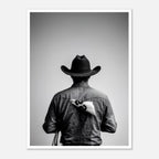 Black and white photograph of a rodeo cowboy viewed from behind wearing a western hat with a numbered rodeo tag pinned to his shirt after competition.