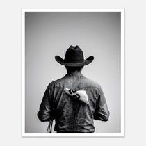 Black and white photograph of a rodeo cowboy viewed from behind wearing a western hat with a numbered rodeo tag pinned to his shirt after competition.