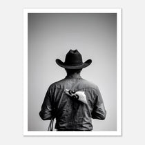 Black and white photograph of a rodeo cowboy viewed from behind wearing a western hat with a numbered rodeo tag pinned to his shirt after competition.
