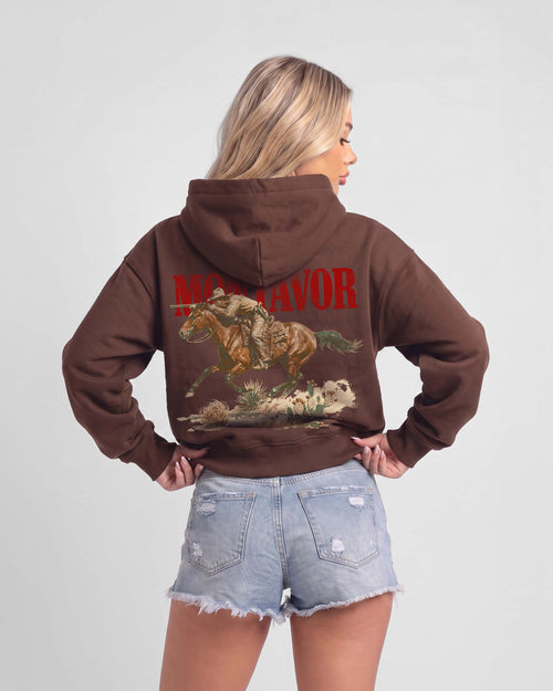A woman shown from the back wearing a brown pullover hoodie with a large graphic print of a cowboy riding a horse and the word 'MONTAVOR' in red, paired with light blue denim shorts against a light gray studio background.