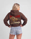 A woman shown from the back wearing a brown pullover hoodie with a large graphic print of a cowboy riding a horse and the word 'MONTAVOR' in red, paired with light blue denim shorts against a light gray studio background.