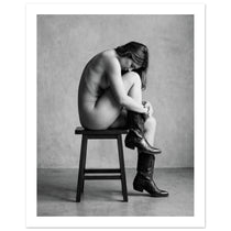 Black and white fine art photograph of a woman seated on a stool wearing cowboy boots in a minimalist studio setting.