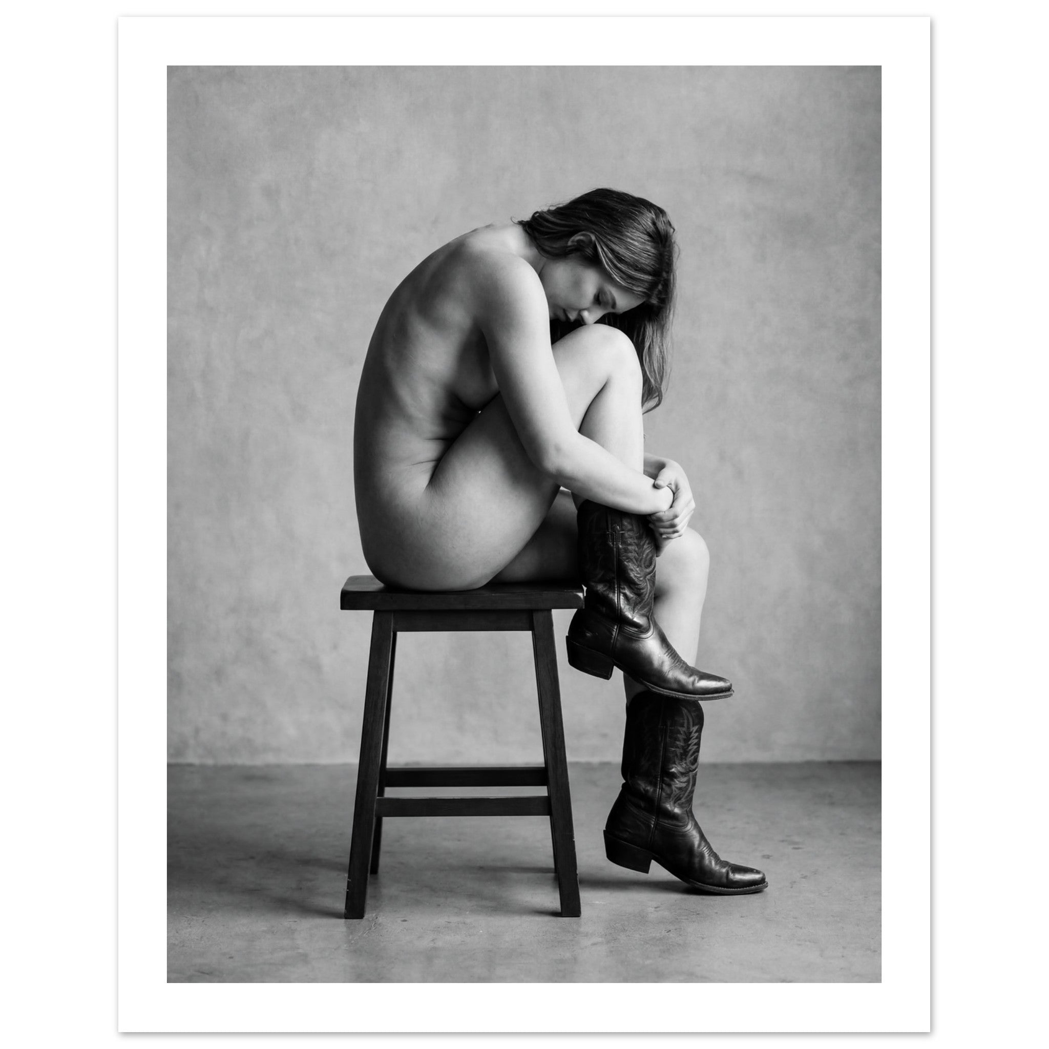 Black and white fine art photograph of a woman seated on a stool wearing cowboy boots in a minimalist studio setting.