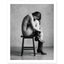 Black and white fine art photograph of a woman seated on a stool wearing cowboy boots in a minimalist studio setting.