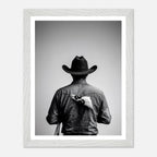 Black and white photograph of a rodeo cowboy viewed from behind wearing a western hat with a numbered rodeo tag pinned to his shirt after competition.