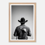 Black and white photograph of a rodeo cowboy viewed from behind wearing a western hat with a numbered rodeo tag pinned to his shirt after competition.