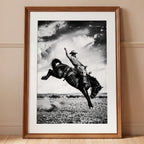 Framed black and white photograph of a cowboy riding a bucking horse against a cloudy sky.