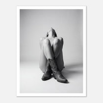 Black and white fine art poster of a seated cowgirl figure wearing classic cowboy boots against a minimalist studio background, creating a dramatic western-inspired composition.