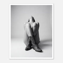 Black and white fine art poster of a seated cowgirl figure wearing classic cowboy boots against a minimalist studio background, creating a dramatic western-inspired composition.