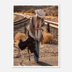 Cowgirl walking through desert carrying a saddle near railroad tracks, western wall art.
