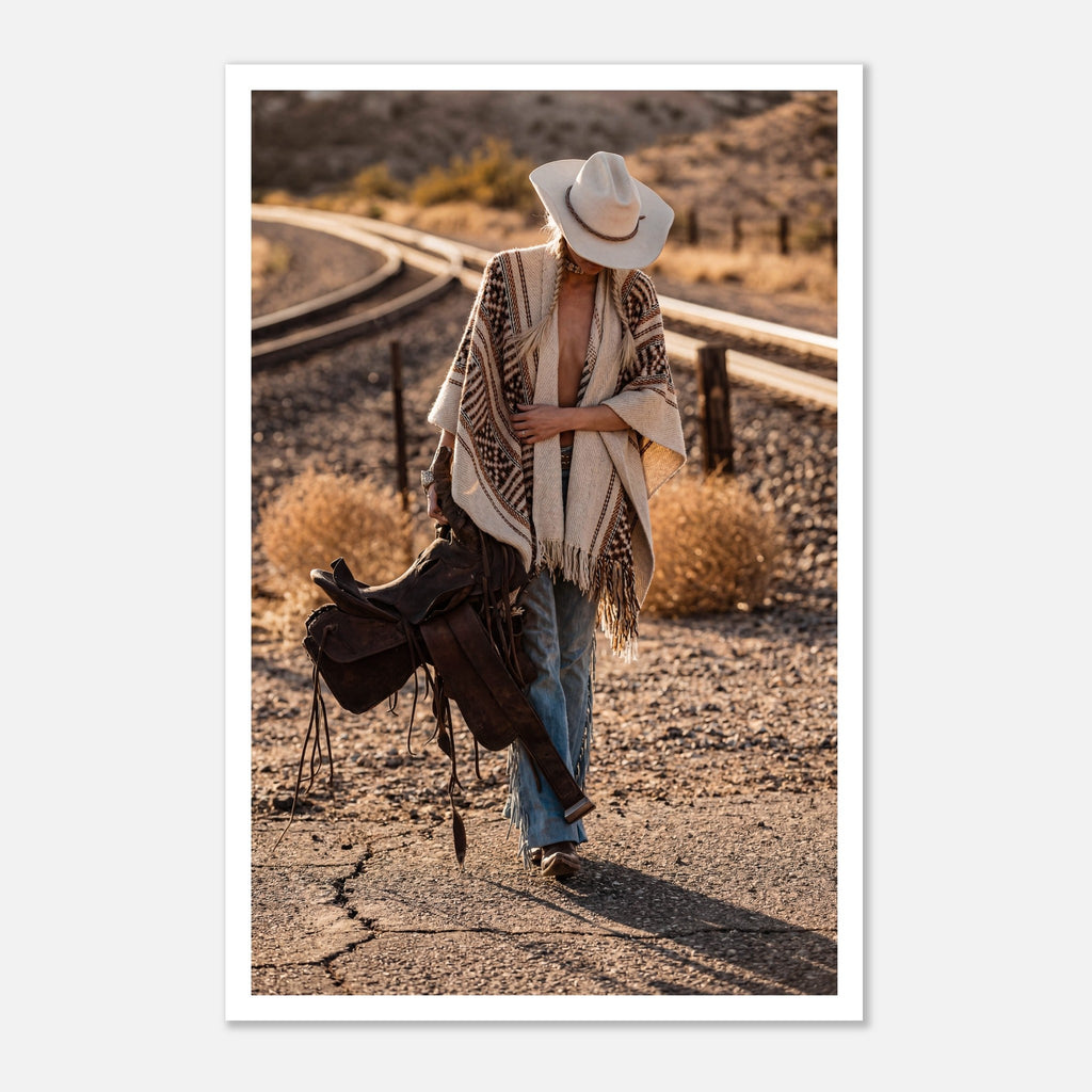 Cowgirl walking through desert carrying a saddle near railroad tracks, western wall art.