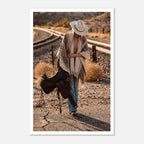 Cowgirl walking through desert carrying a saddle near railroad tracks, western wall art.