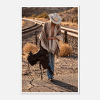 Cowgirl walking through desert carrying a saddle near railroad tracks, western wall art.