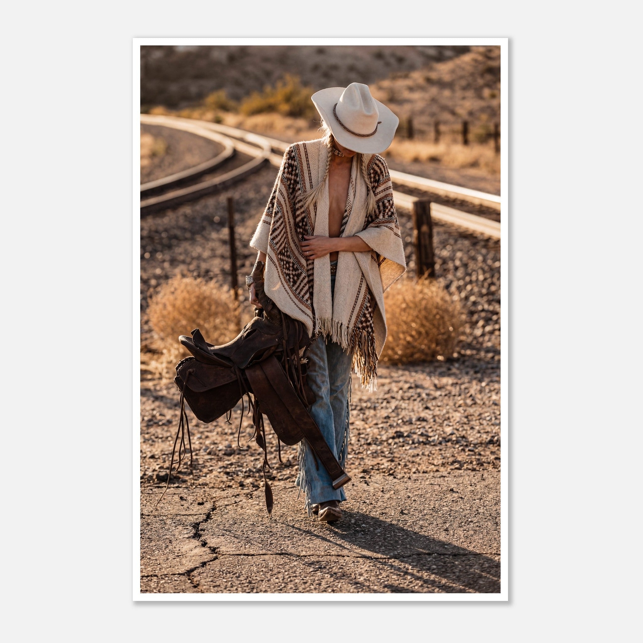 Cowgirl walking through desert carrying a saddle near railroad tracks, western wall art.