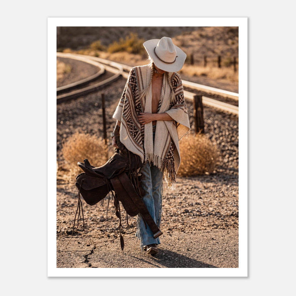 Cowgirl walking through desert carrying a saddle near railroad tracks, western wall art.