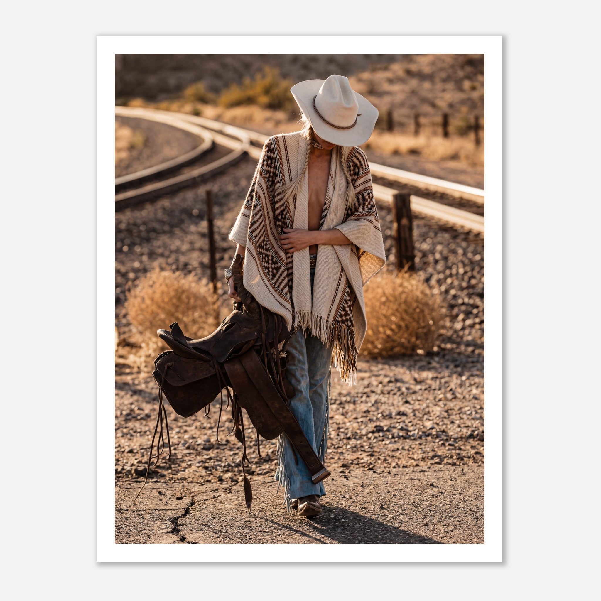 Cowgirl walking through desert carrying a saddle near railroad tracks, western wall art.