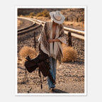 Cowgirl walking through desert carrying a saddle near railroad tracks, western wall art.