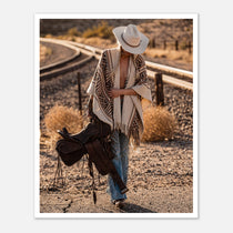 Cowgirl walking through desert carrying a saddle near railroad tracks, western wall art.