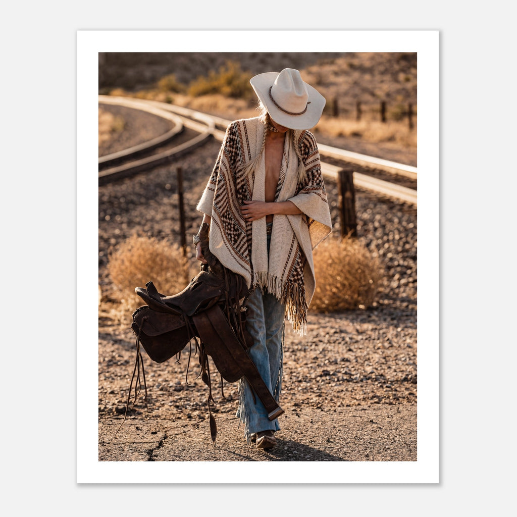 Cowgirl walking through desert carrying a saddle near railroad tracks, western wall art.