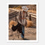 Cowgirl walking through desert carrying a saddle near railroad tracks, western wall art.